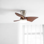 Close view of a Clérachic ceiling fan in nickel with walnut-grain blades and a white light, with sheer curtains behind.