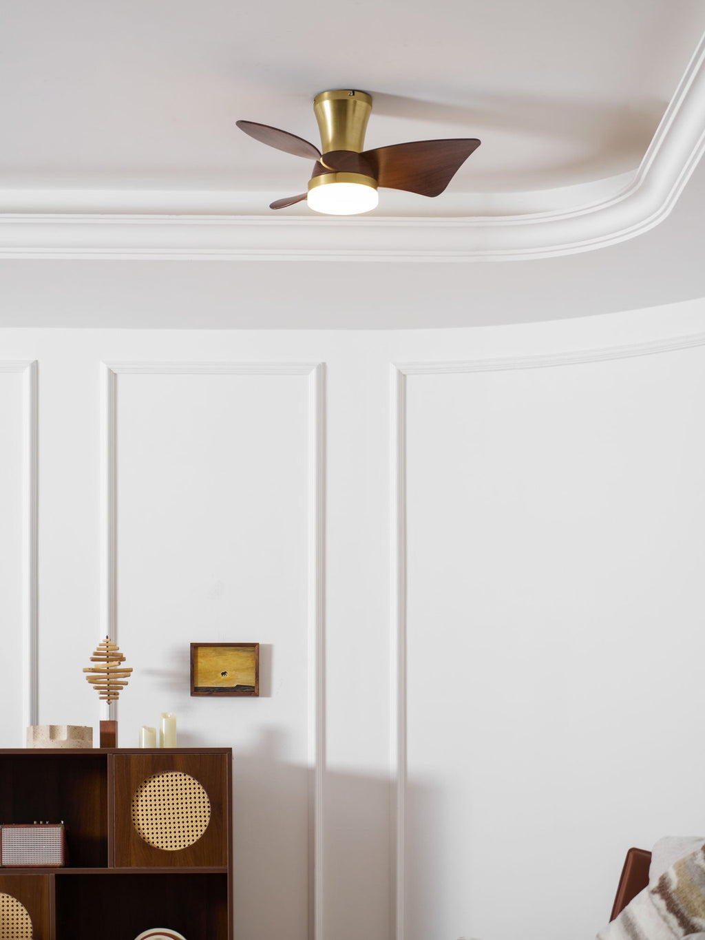 A Clérachic gold ceiling fan with its light on, high above a white wall with a framed picture and a cane-fronted cabinet.