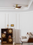 A Clérachic gold ceiling fan with a lit light, hung over a white room with ornate ceiling moulding and a cabinet with cane doors.
