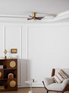 A Clérachic gold ceiling fan high on a white ceiling, seen from far below, with a wooden cabinet and armchair at the foot of the wall.