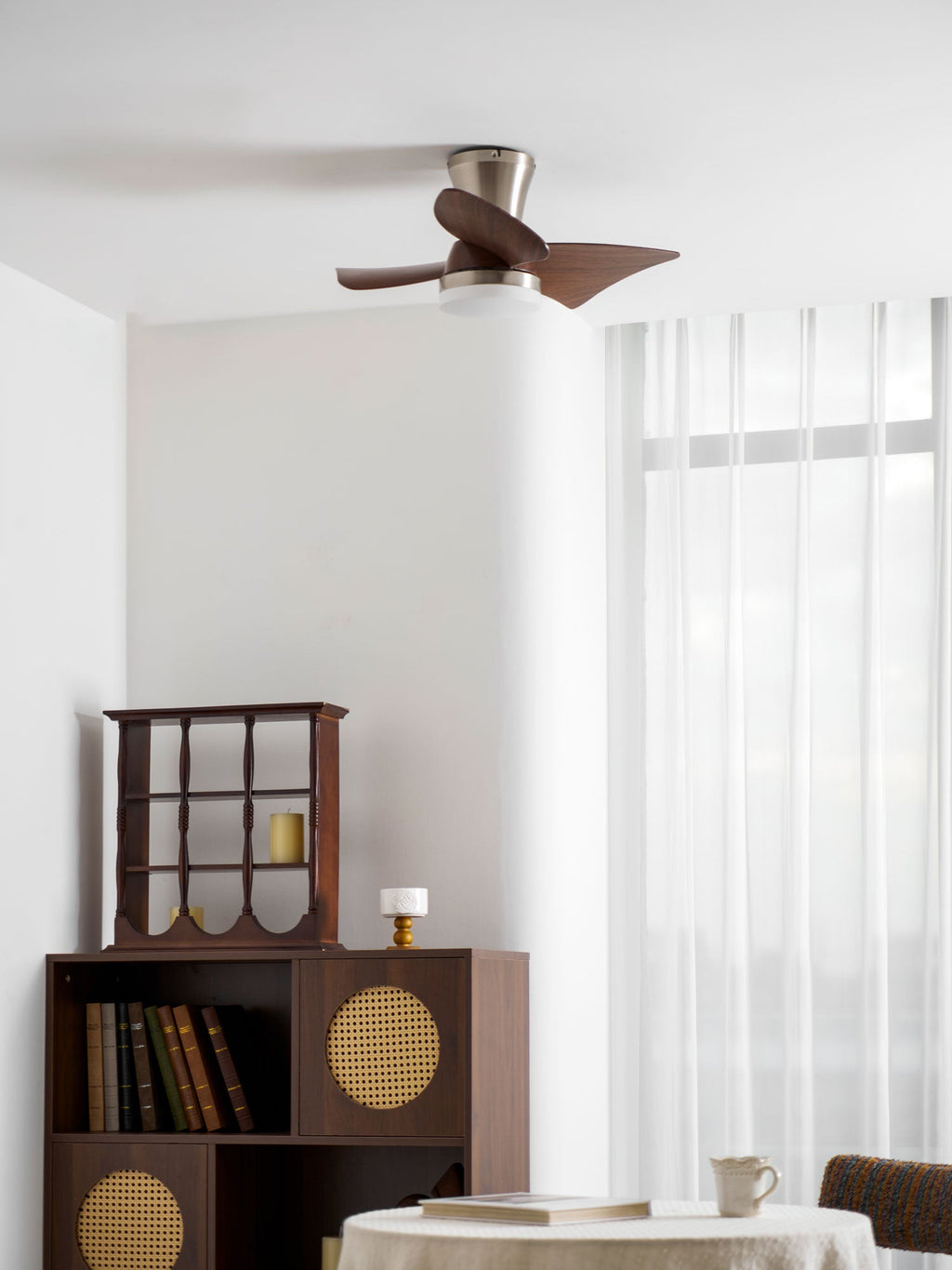 The Clérachic nickel ceiling fan viewed from the side, over a bookcase and a cane-doored cabinet in front of sheer white curtains.