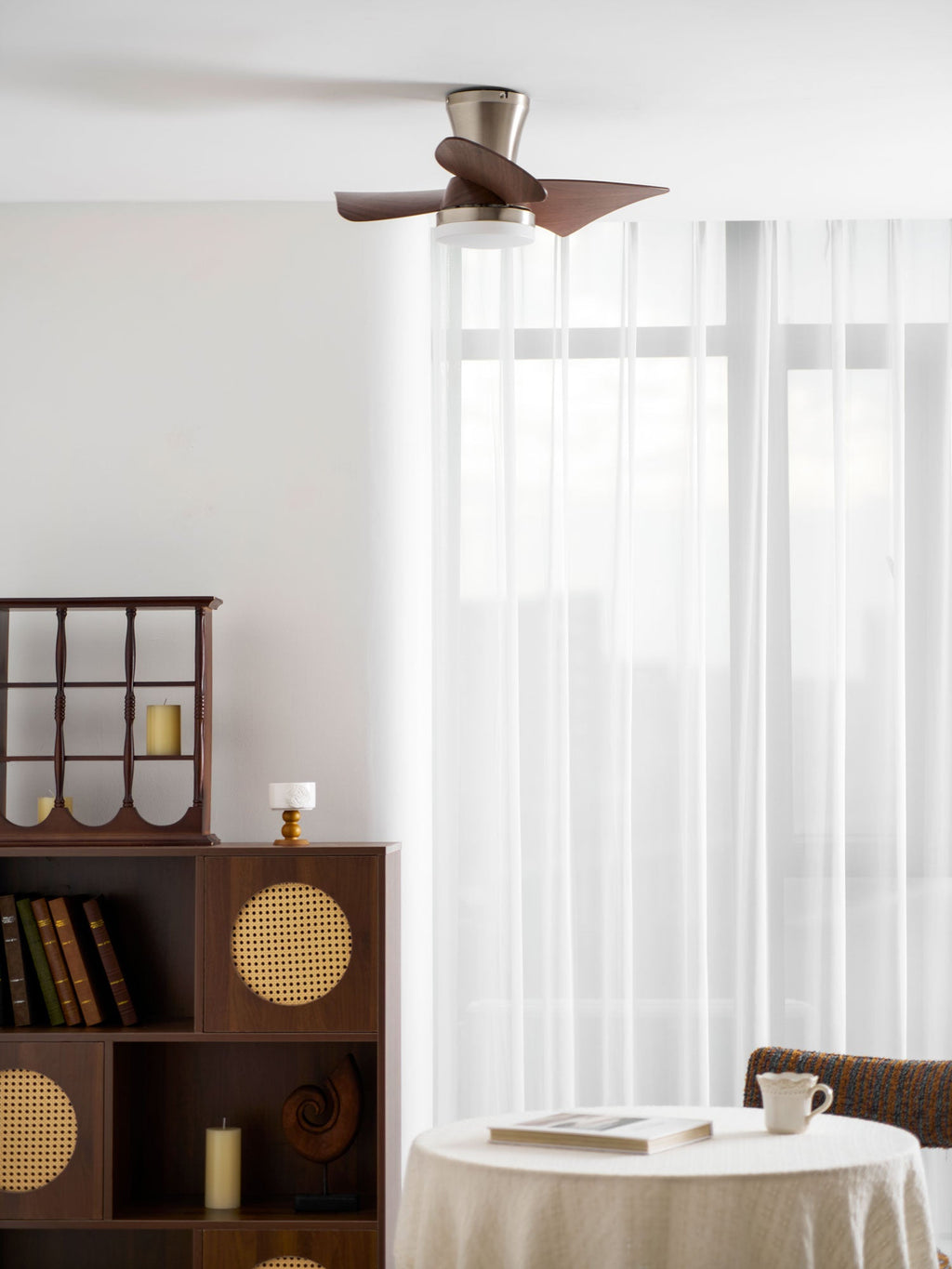 A Clérachic nickel ceiling fan above a tall display cabinet, a bookshelf and a table with a teacup, with light curtains at the window.