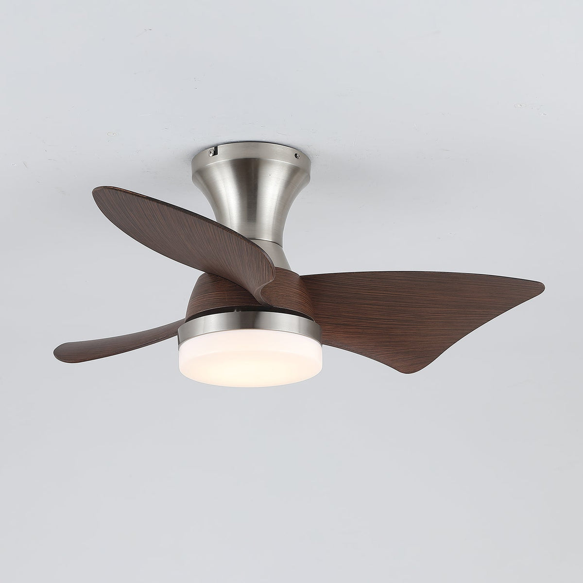 A Clérachic nickel ceiling fan from the side, with a warm glowing light and the blades swept around its slim trumpet-shaped canopy.