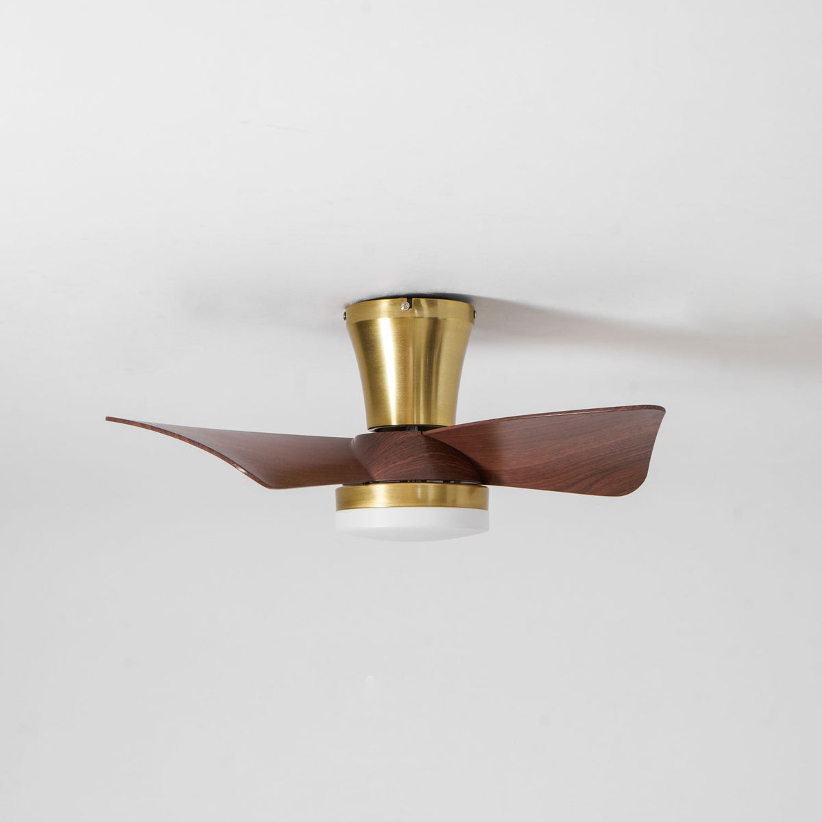 A low-angle view of the Clérachic gold ceiling fan, with its trumpet canopy, gold trim ring and unlit light under walnut-grain blades.