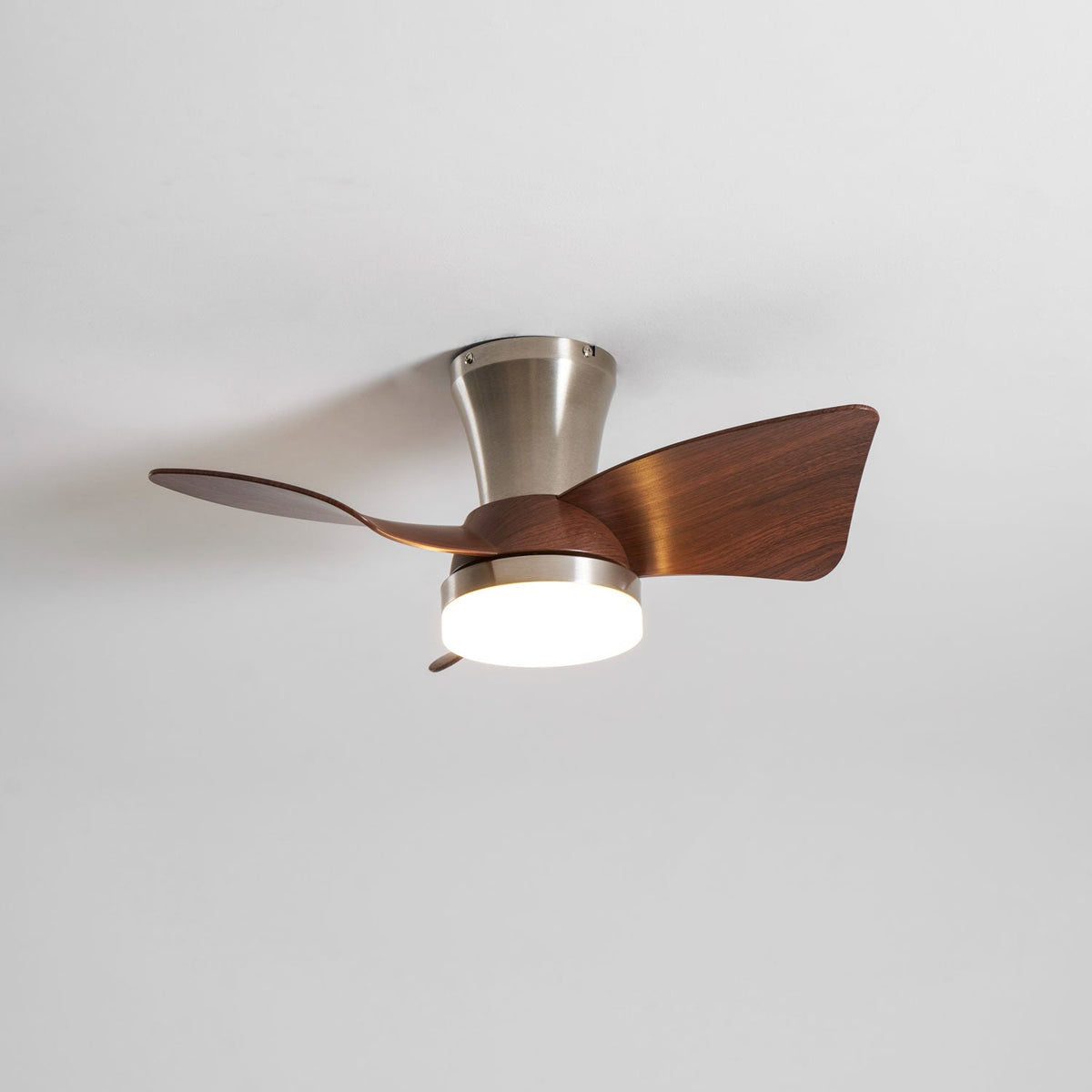 A Clérachic nickel ceiling fan with a warm light on, shown from below with its blades turning against a white ceiling.