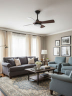 Black-stemmed Clérachic fan with walnut blades above a living room with a grey sofa, blue armchairs and a patterned rug.