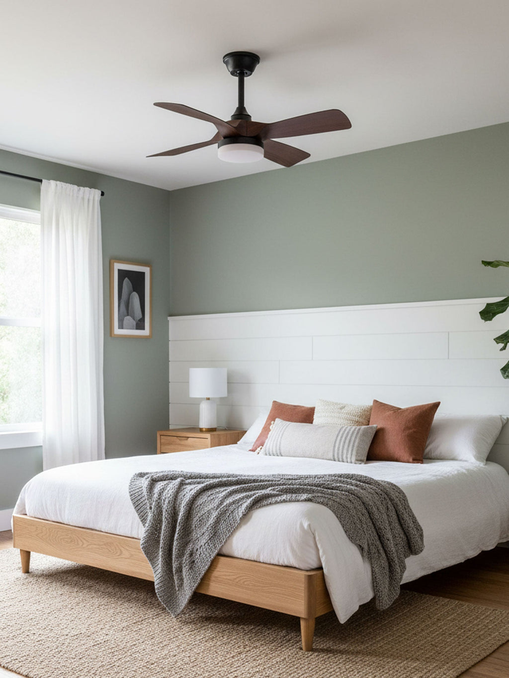 Walnut-finish fan blades turn above a bed with white linen and a woven rug, in a bedroom with a pale sage wall, from Clérachic.