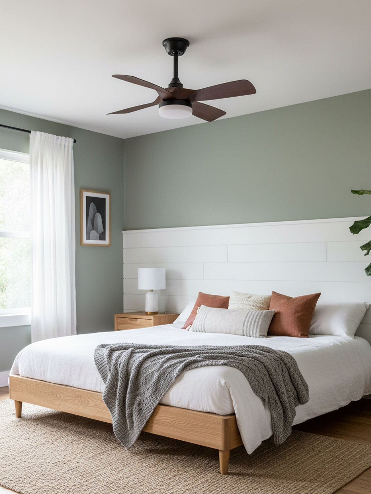 Walnut-finish fan blades turn above a bed with white linen and a woven rug, in a bedroom with a pale sage wall, from Clérachic.