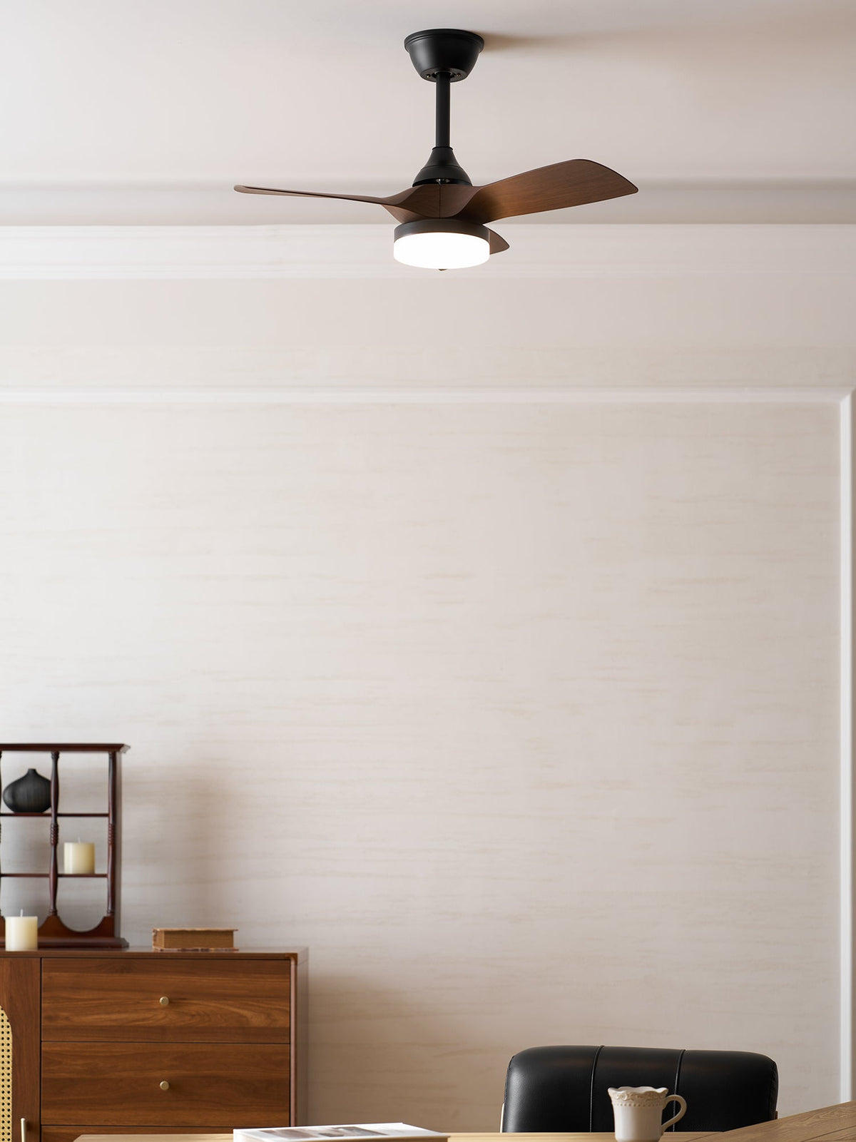 This Clérachic ceiling fan, with its light on, hangs in a calm cream room with a wooden sideboard and a black leather chair.