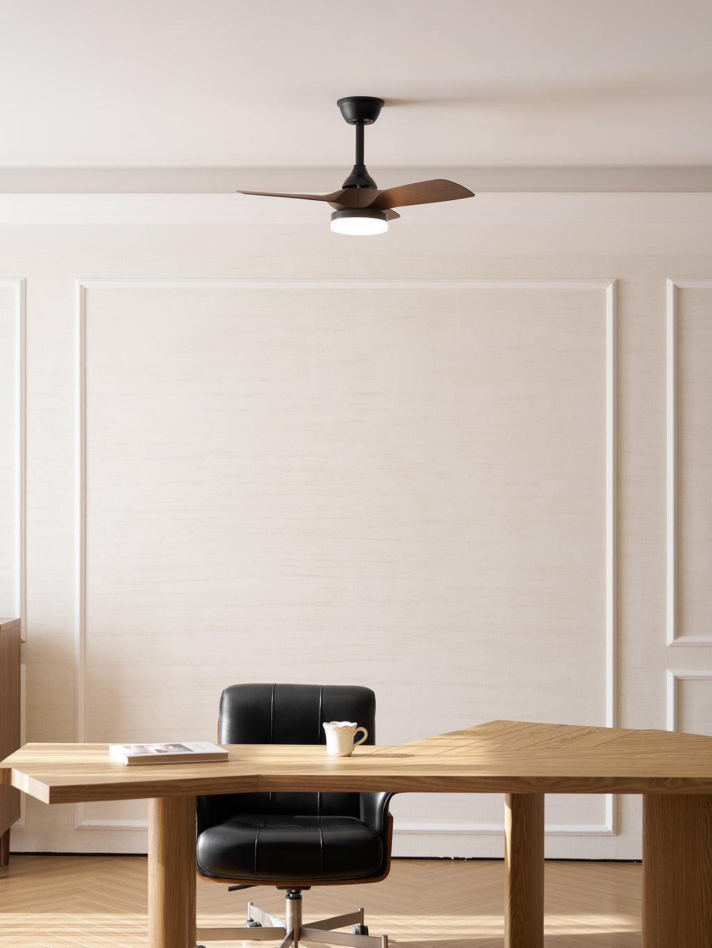 This Clérachic fan hangs in front of a panelled wall, above a pale oak desk with a mug and a black office chair.
