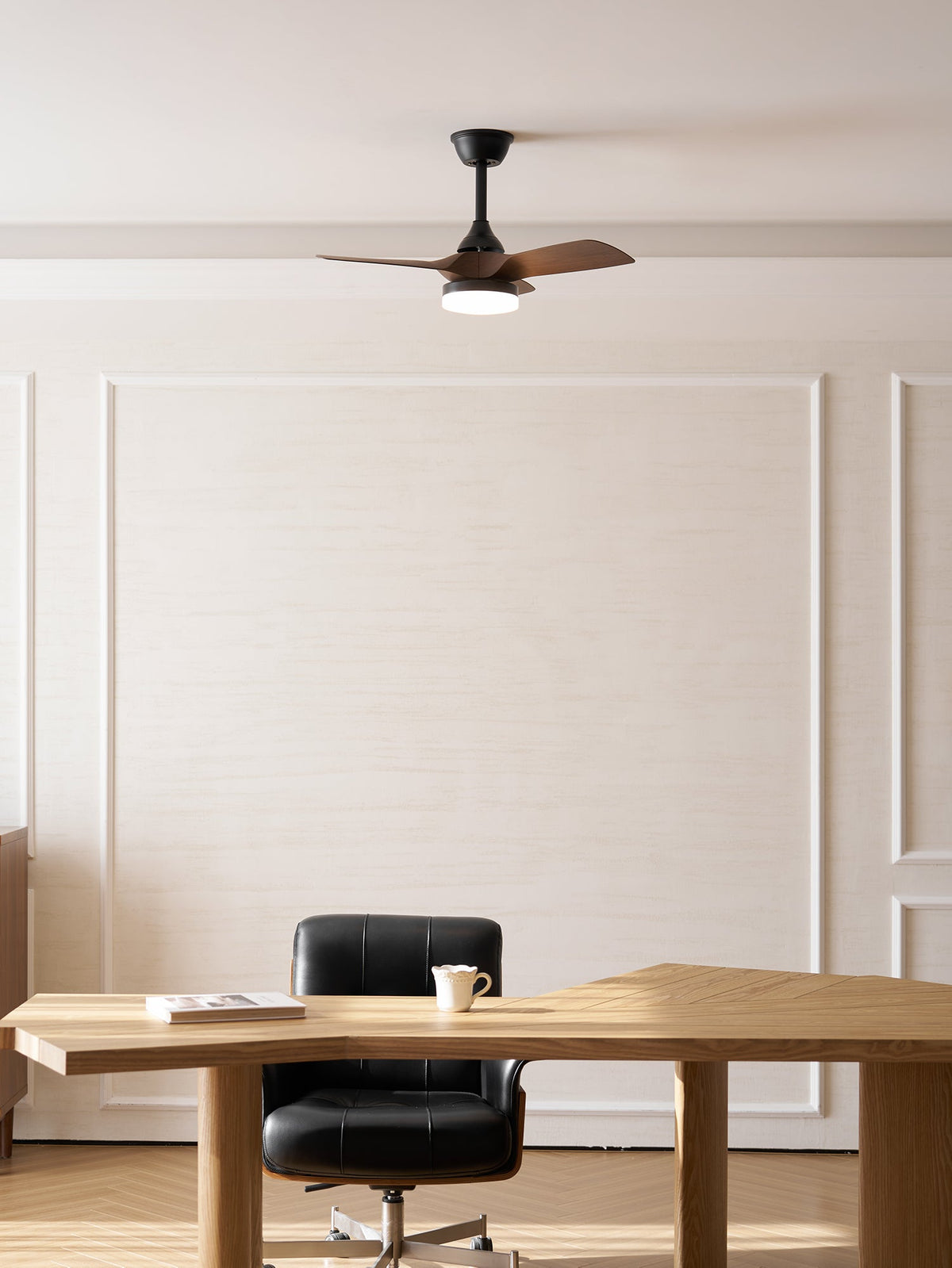 This Clérachic fan hangs in front of a panelled wall, above a pale oak desk with a mug and a black office chair.