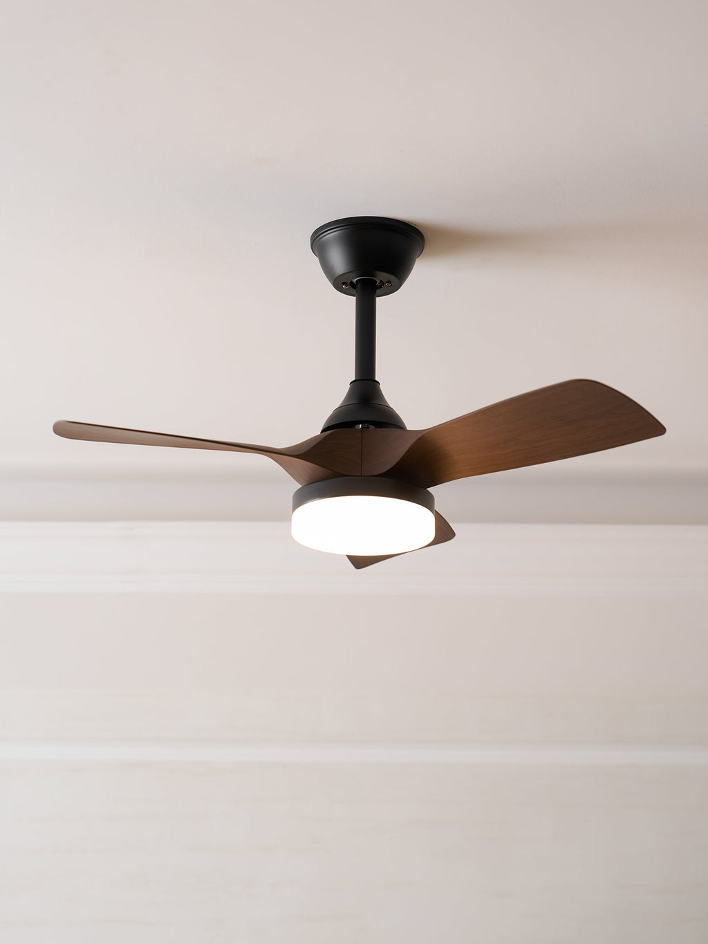 Looking up at this Clérachic ceiling fan with its light on, its black stem and walnut-grain blades against a pale ceiling.