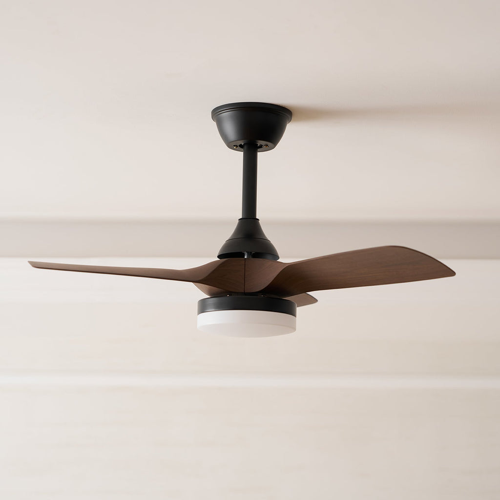 Side-on view of this Clérachic fan, with its black canopy, flat walnut-grain blades and white light cover, against a cream ceiling.