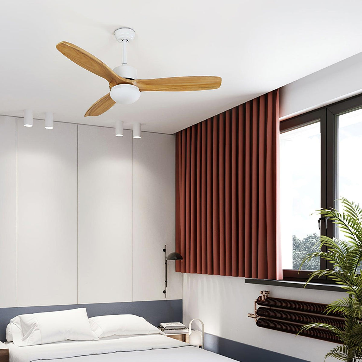 White ceiling fan from Clérachic with wooden blades over a bedroom with a rust-red curtain, spotlights and a large window.