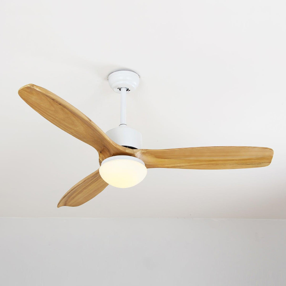 Ceiling fan from Clérachic with wooden blades and its round light switched on, below a pale ceiling.