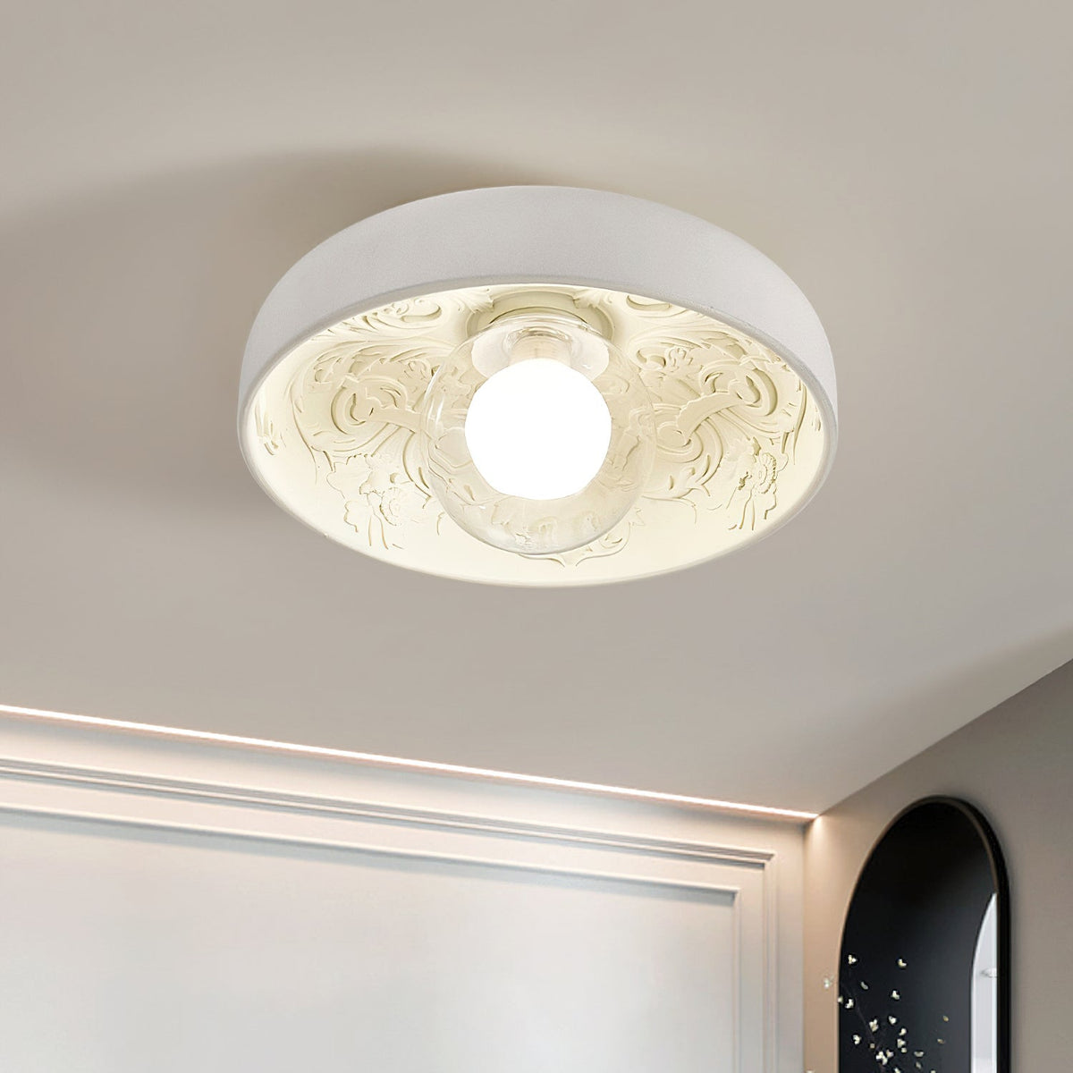 Close view of the Clérachic bowl light's carved scrollwork and glowing globe, with a cove light line along the wall behind.