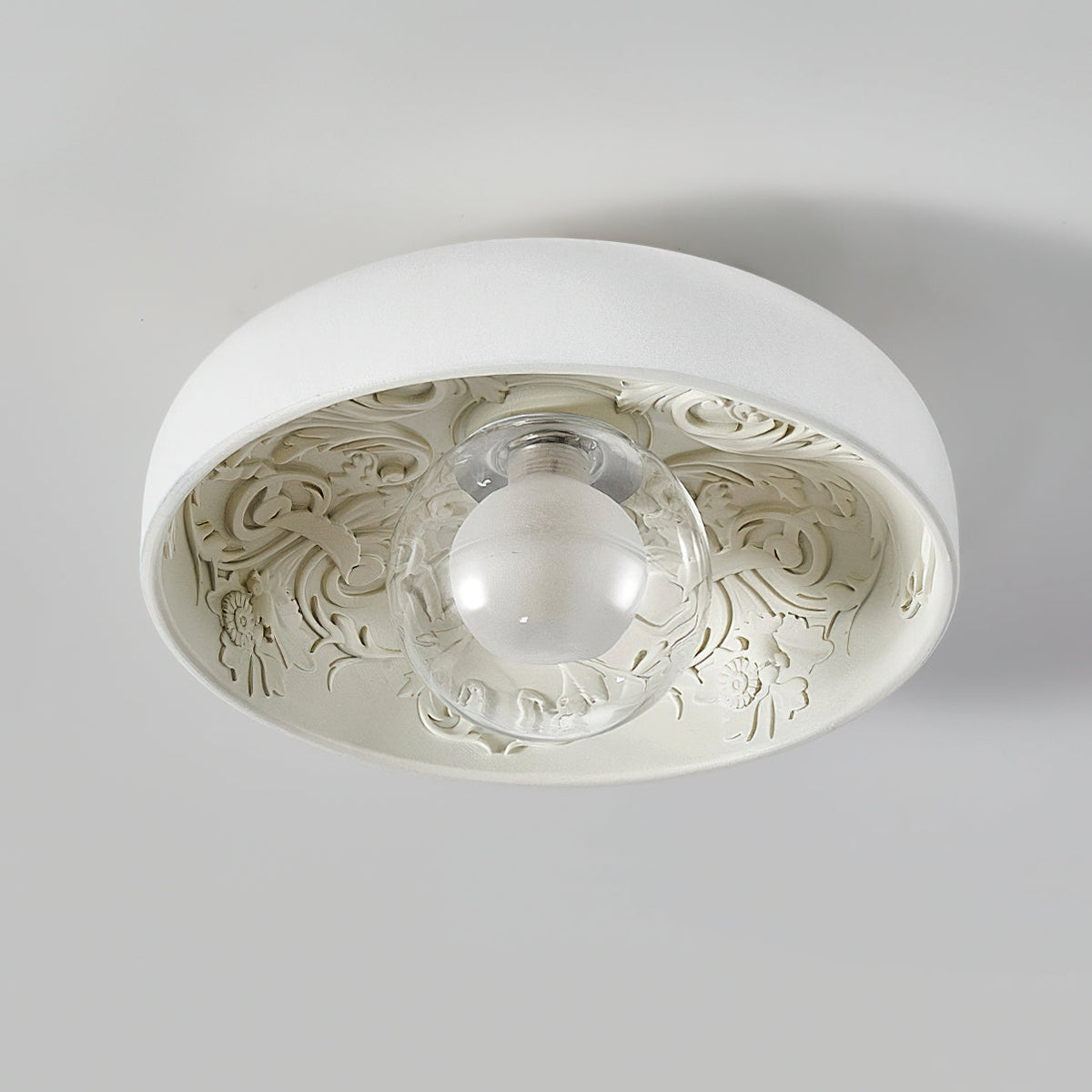 This Clérachic ceiling light with a clear glass globe, switched off, shows its carved floral pattern on a pale grey ceiling.
