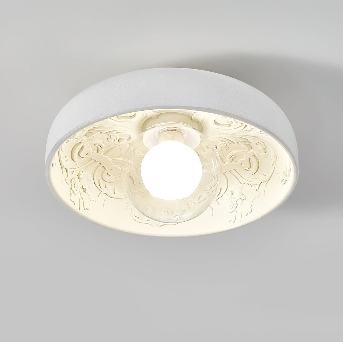 Clérachic bowl light lit with a clear glass globe, its carved interior glowing warm cream against a pale grey ceiling.