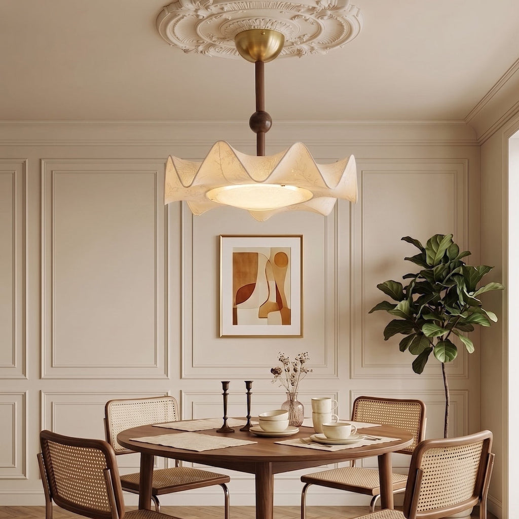 A Clérachic ceiling light hangs over a round dining table laid with cups, in front of a framed print and a large plant.