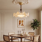 A Clérachic ceiling light hangs over a round dining table laid with cups, in front of a framed print and a large plant.