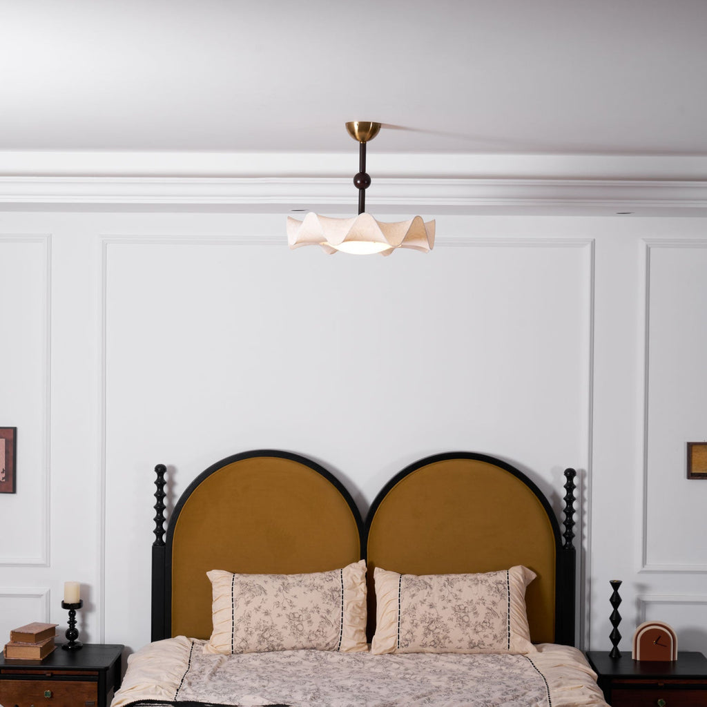 A small Clérachic ceiling light seen above a bed with a rounded mustard headboard and floral pillows, in a white-panelled room.