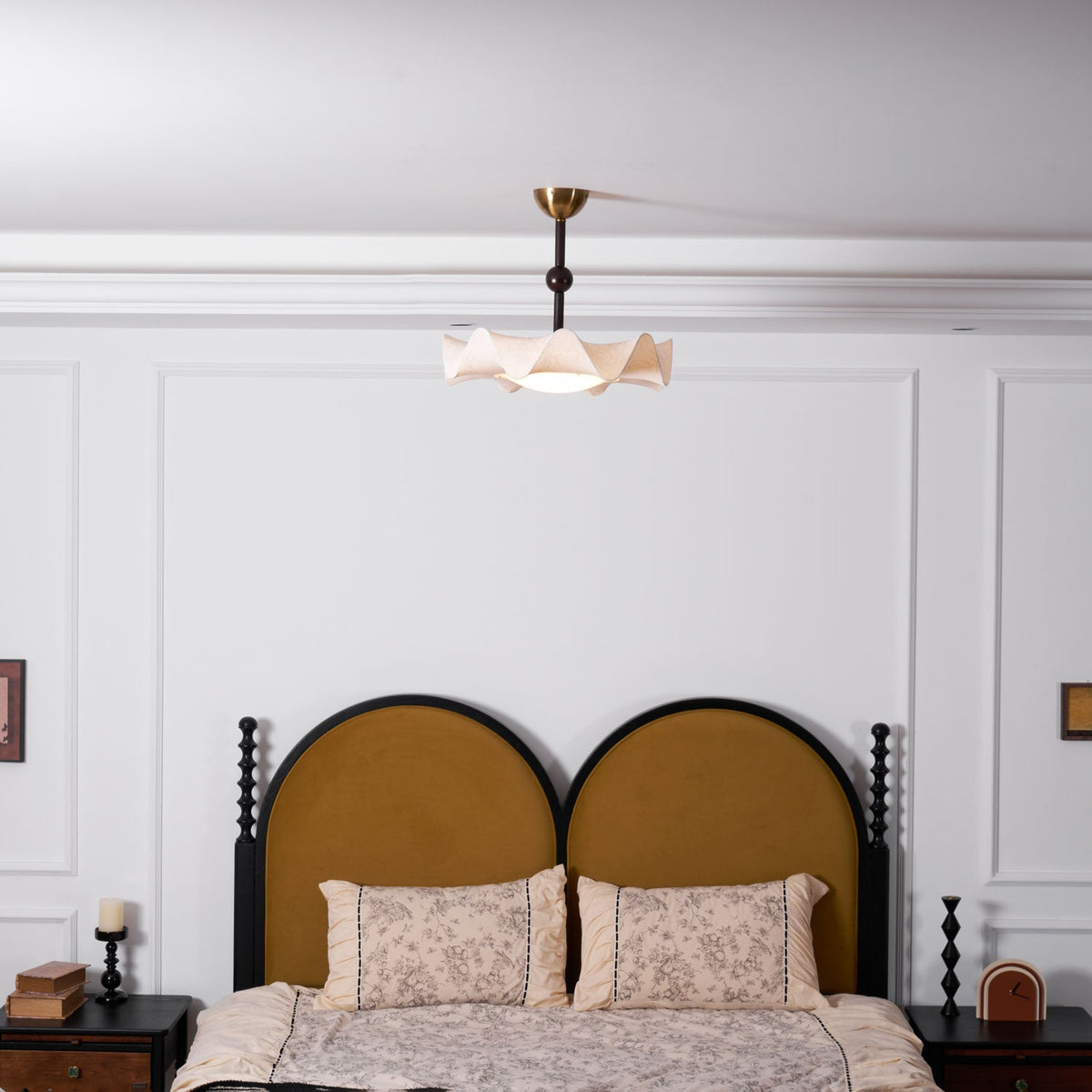 A small Clérachic ceiling light seen above a bed with a rounded mustard headboard and floral pillows, in a white-panelled room.