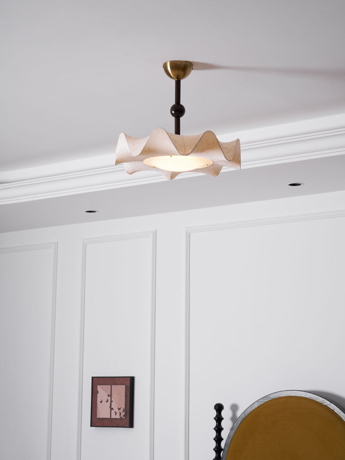 A Clérachic ceiling light lit above a white-panelled bedroom wall, with a corner of the bed and a bedside table below.