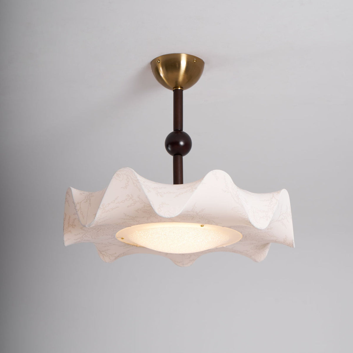Lit Clérachic ceiling light against a grey ceiling, showing the brass canopy, slim stem and a warm glowing diffuser.