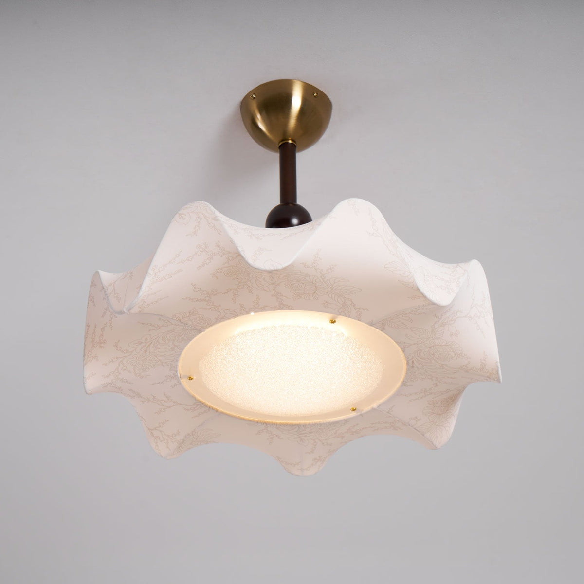 Straight view of this lit Clérachic ceiling light, with a brass canopy, a walnut-toned stem and a ruffled shade around the glow.