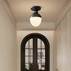 A black-topped ceiling light with a frosted white globe glows in a hallway below cornice moulding, facing an arched black-framed glazed door, from Clérachic.