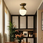 Lit white glass globe with an antique brass top on this Clérachic ceiling light, above a dining corner with a leafy plant and orange flowers.