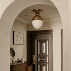 Beneath a cream arched ceiling, a brass-topped Clérachic ceiling light with a white globe sits above a panelled wooden door and a small console.