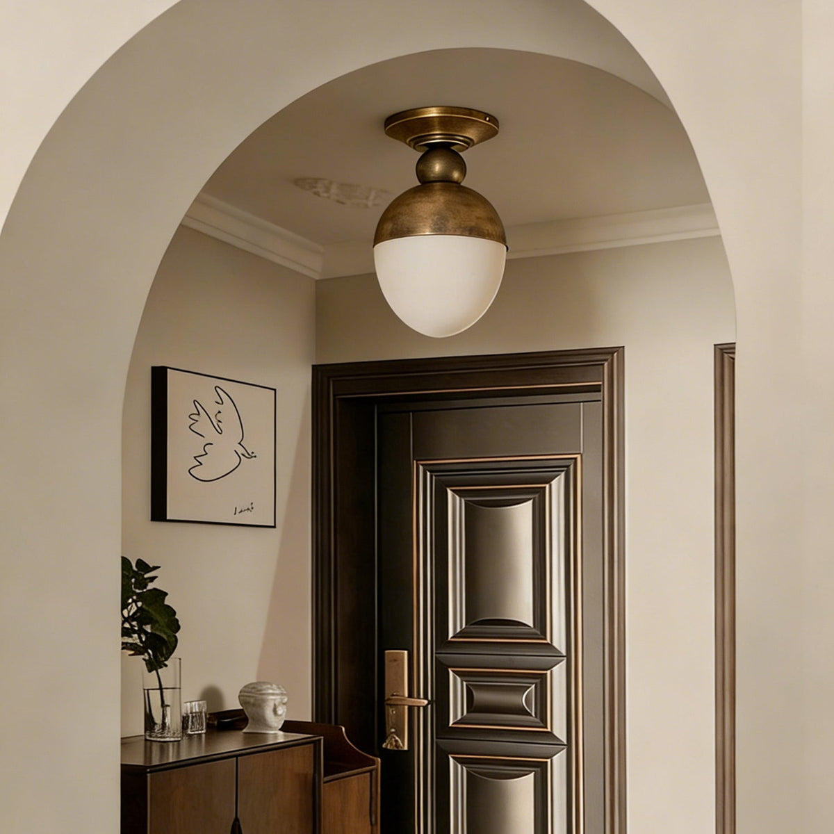 Beneath a cream arched ceiling, a brass-topped Clérachic ceiling light with a white globe sits above a panelled wooden door and a small console.