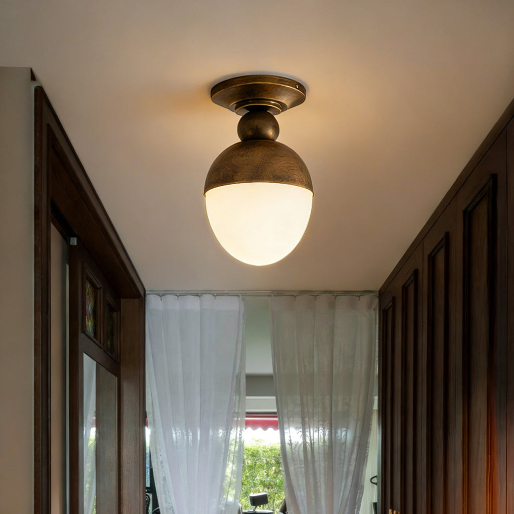 Brass cap and lit opal globe seen from below in a dark wood corridor, this Clérachic ceiling light has sheer white curtains at the far end.