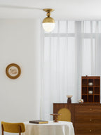 A small brass-topped Clérachic ceiling light hangs high in a bright room with sheer curtains, a wooden sideboard and a round table at the foot of the frame.