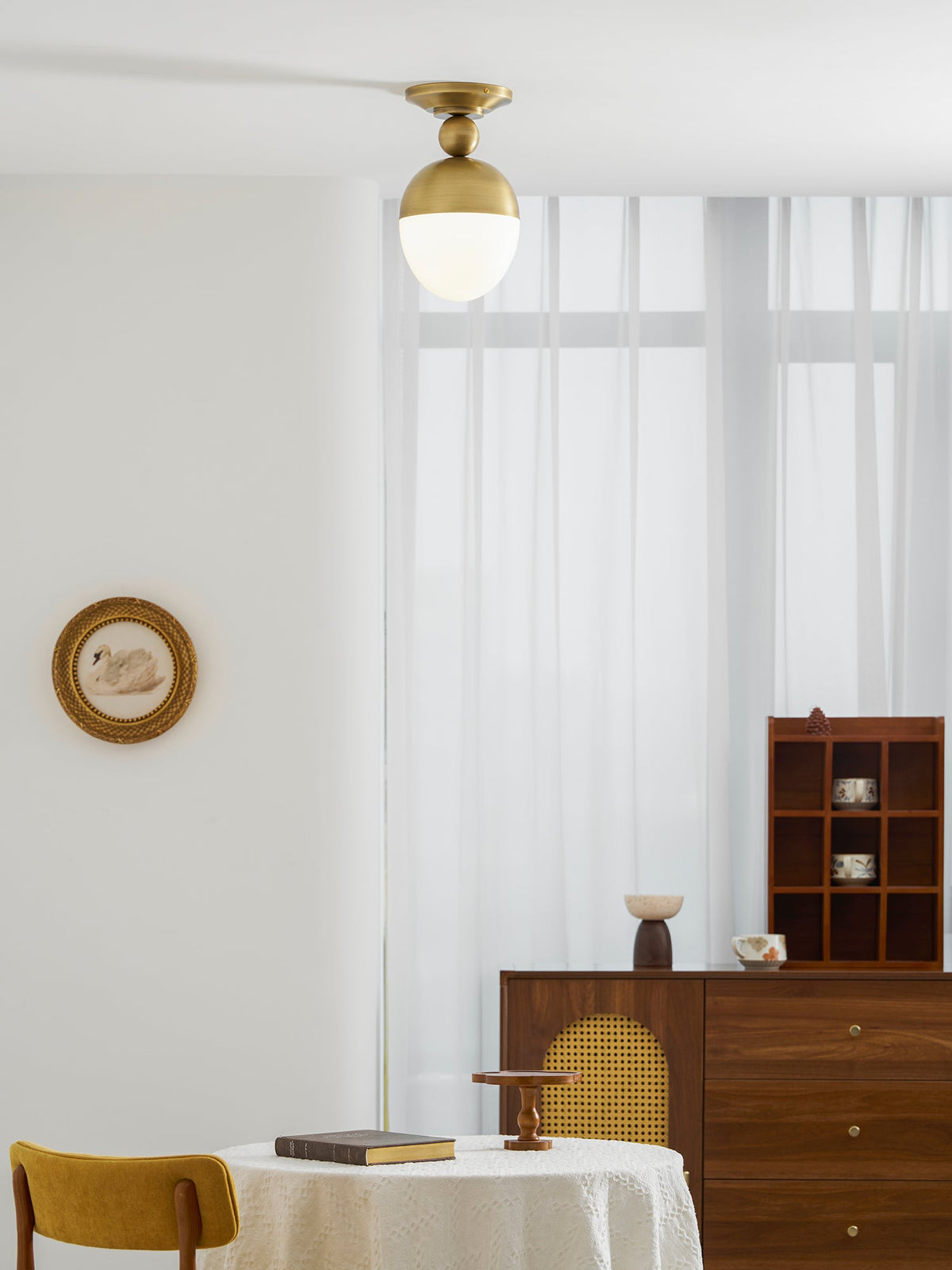 A small brass-topped Clérachic ceiling light hangs high in a bright room with sheer curtains, a wooden sideboard and a round table at the foot of the frame.