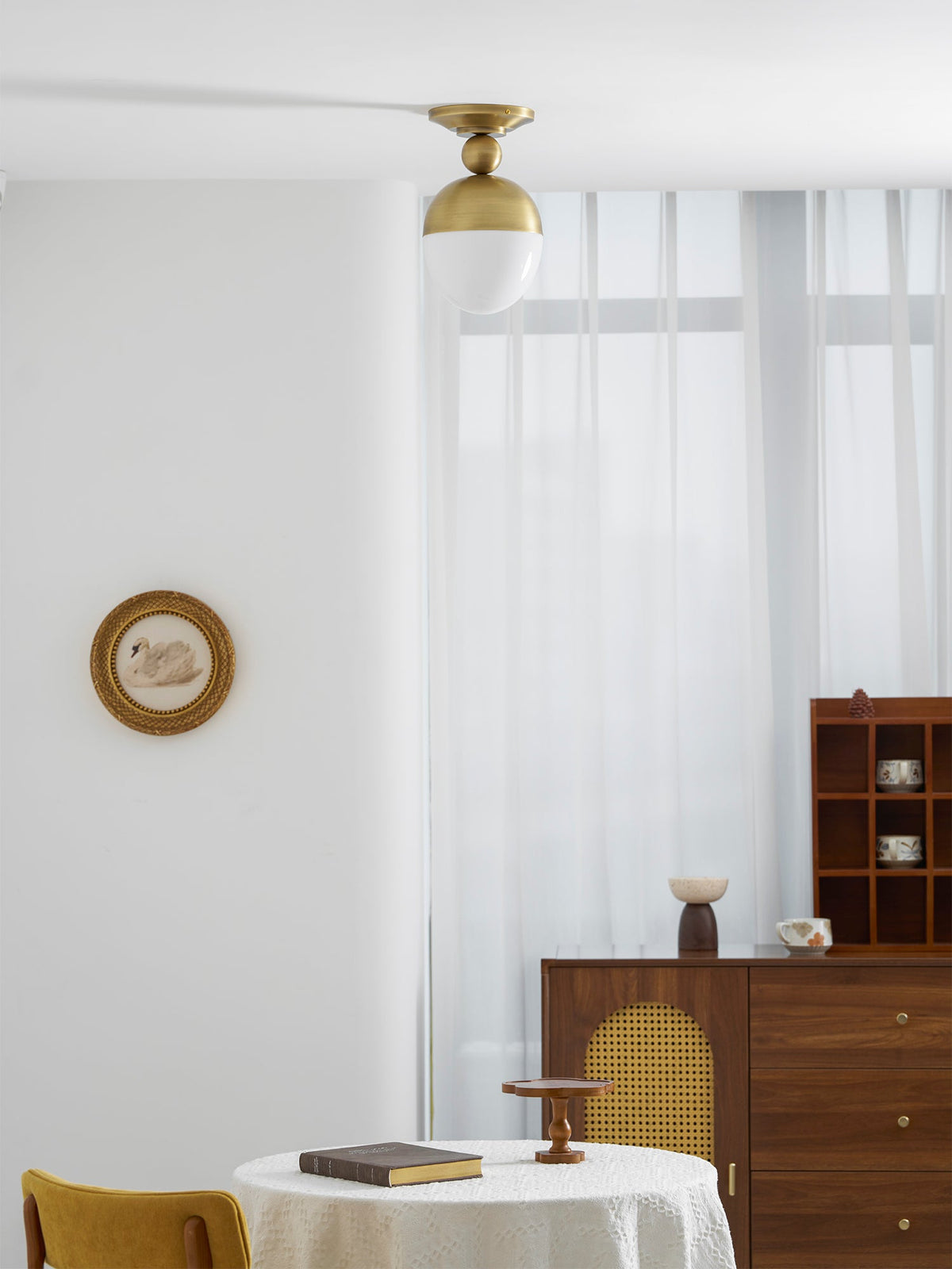 Unlit Clérachic ceiling light with a brass top and white glass globe, above a wooden sideboard with a cane-panel door and sheer curtains.