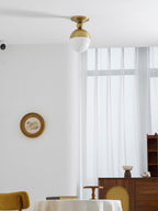 Wider view of the brass-topped Clérachic ceiling light in a pale room, with a mustard chair, a cane-fronted sideboard and a small round picture.
