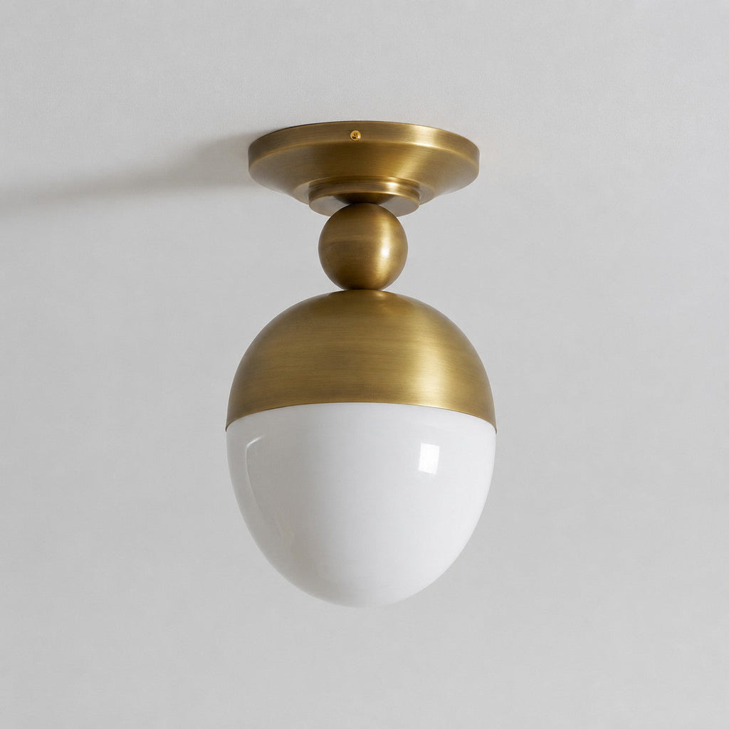 Close, straight-on view of the antique brass canopy, ball joint and unlit white glass globe of this Clérachic ceiling light on a pale ceiling.