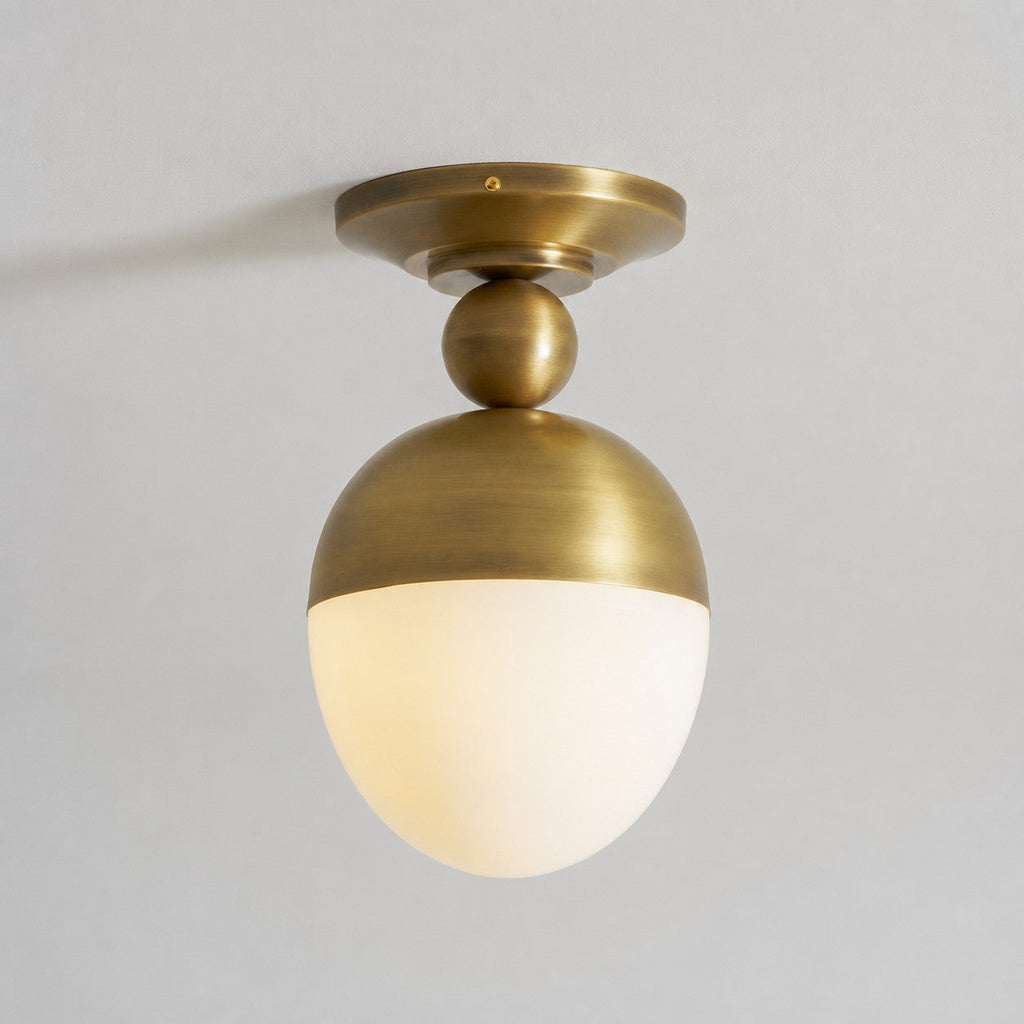 This Clérachic ceiling light with its globe switched on, a soft warm glow beneath the brushed antique brass top, against a pale grey ceiling.
