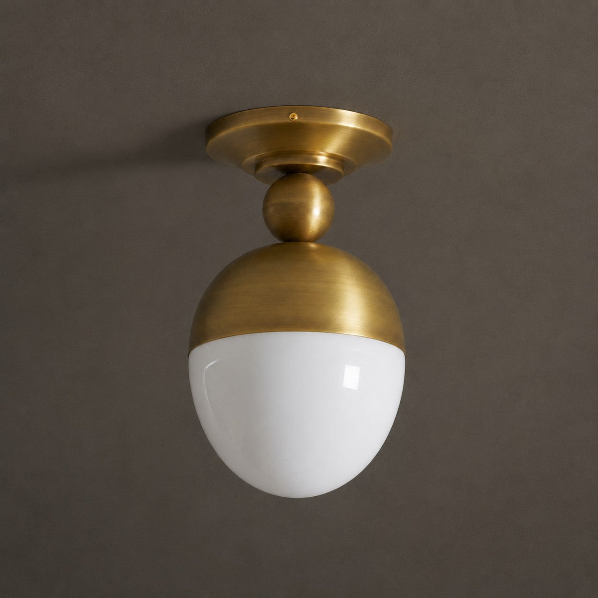 Unlit white glass globe under a brass dome on this Clérachic ceiling light, photographed against a dark taupe backdrop.
