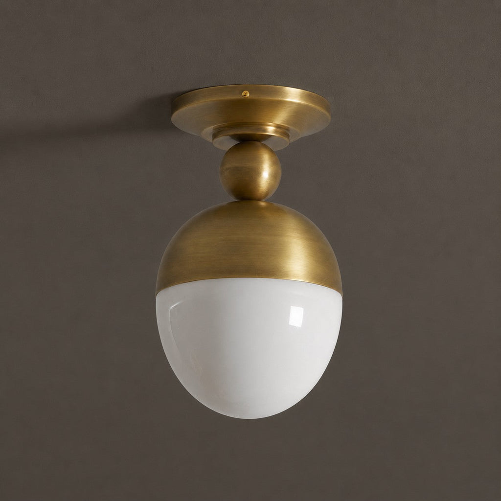 A Clérachic ceiling light in antique brass with a glossy white globe, seen slightly from below against a dark brown-grey wall.