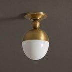 A Clérachic ceiling light in antique brass with a glossy white globe, seen slightly from below against a dark brown-grey wall.