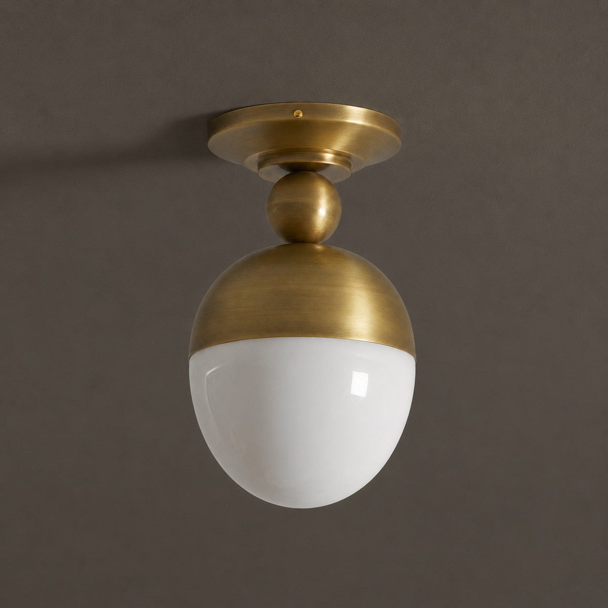 A Clérachic ceiling light in antique brass with a glossy white globe, seen slightly from below against a dark brown-grey wall.