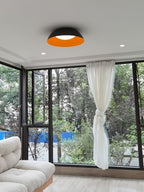 A dark-grey dome ceiling light with an orange inside, from Clérachic, in a room with floor-to-ceiling windows and a knotted curtain.