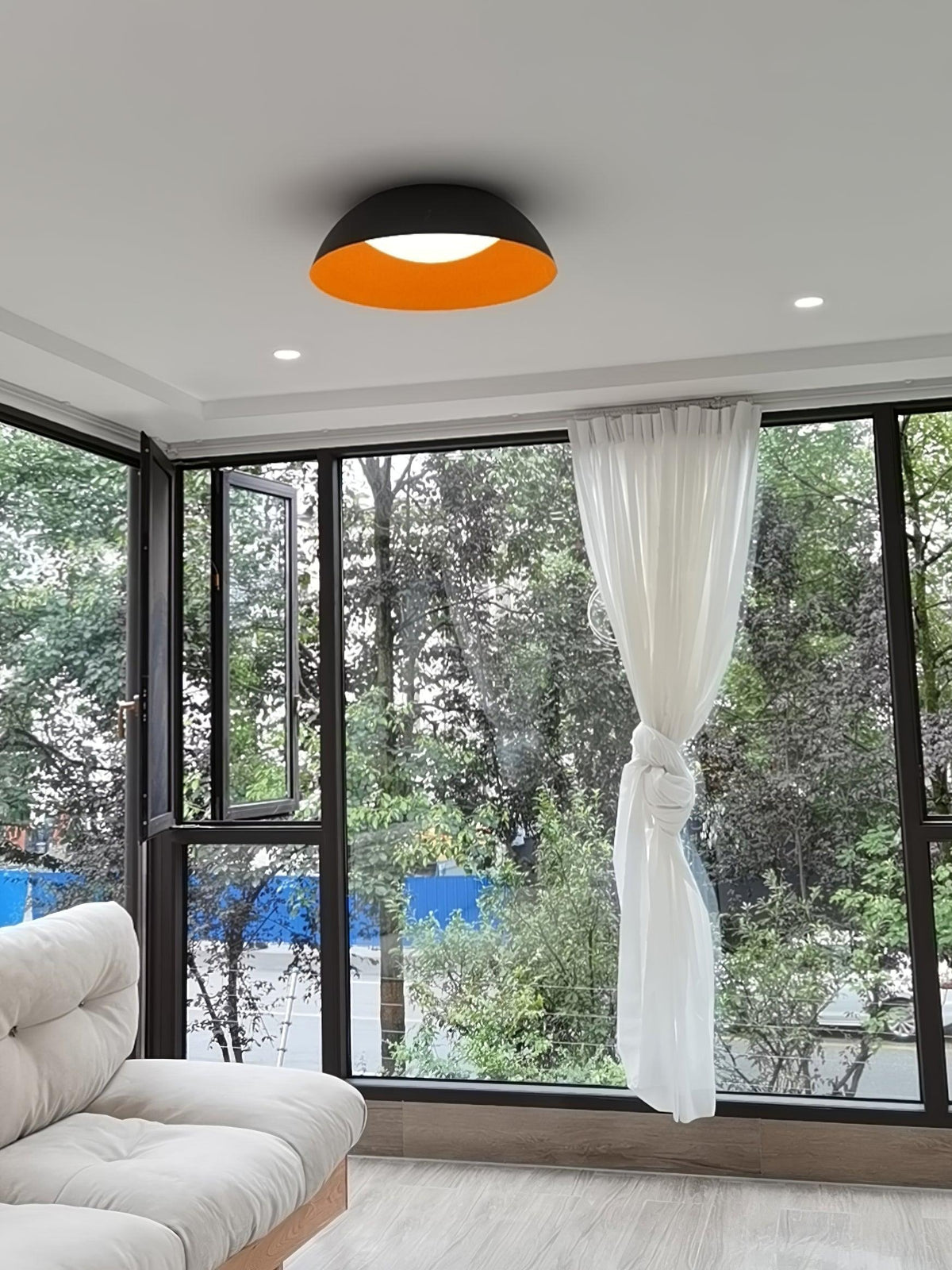 A dark-grey dome ceiling light with an orange inside, from Clérachic, in a room with floor-to-ceiling windows and a knotted curtain.