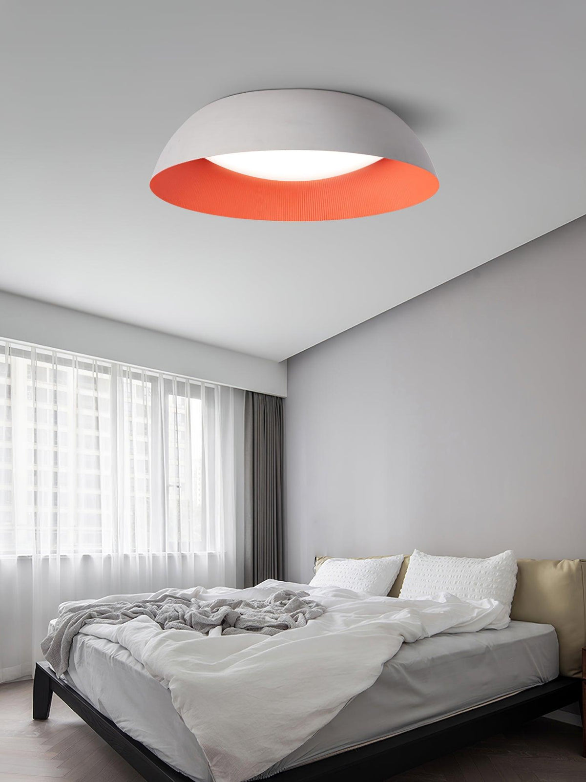 White dome ceiling light with a red-orange inner rim above a grey bedroom with an unmade bed and sheer curtains, from Clérachic.