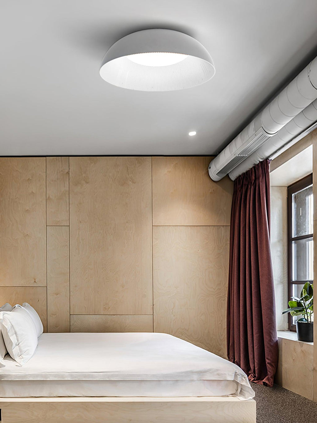 Grey-white dome ceiling light from Clérachic in a bedroom with pale wood panelling, a maroon curtain and an exposed duct.