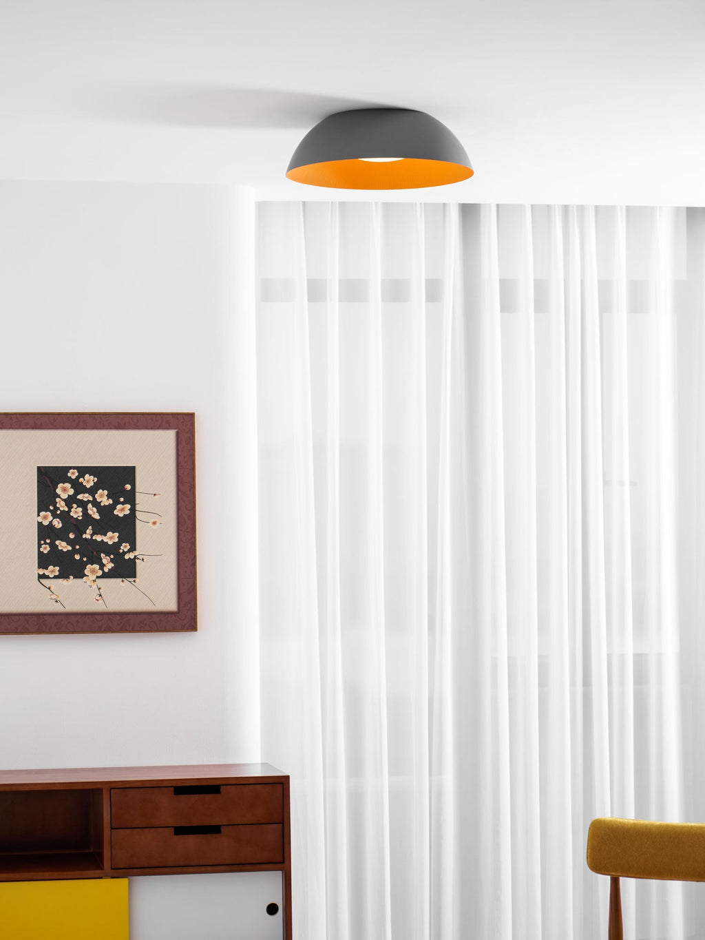 Grey and orange dome ceiling light from Clérachic high above a white room with sheer curtains, a framed print and a sideboard.