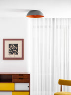Small grey ceiling light with an orange rim from Clérachic seen high in a white room, with a framed print and a yellow-fronted sideboard.